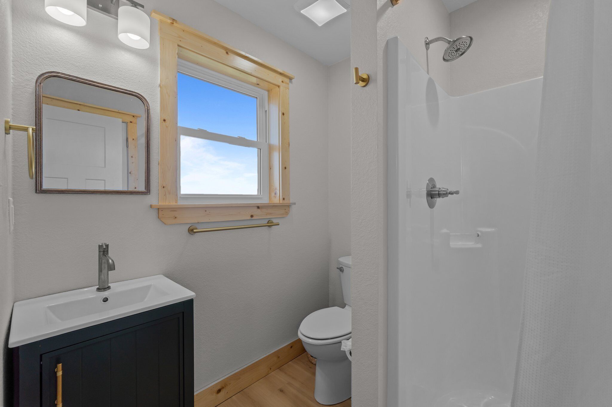 Et badeværelse med en hvid vask, toilet, brusebad med glasdør, trærammevindue og et spejl.