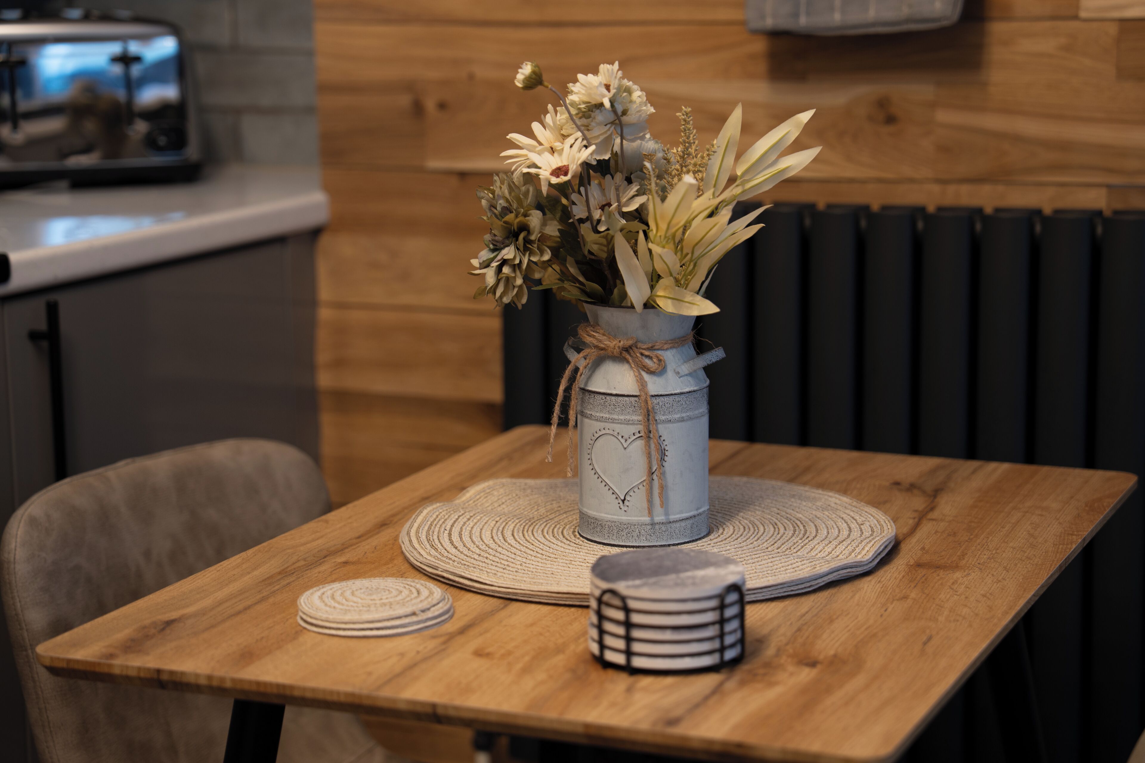 A wooden table with a woven placemat, a vase with flowers, and a stack of coasters.
