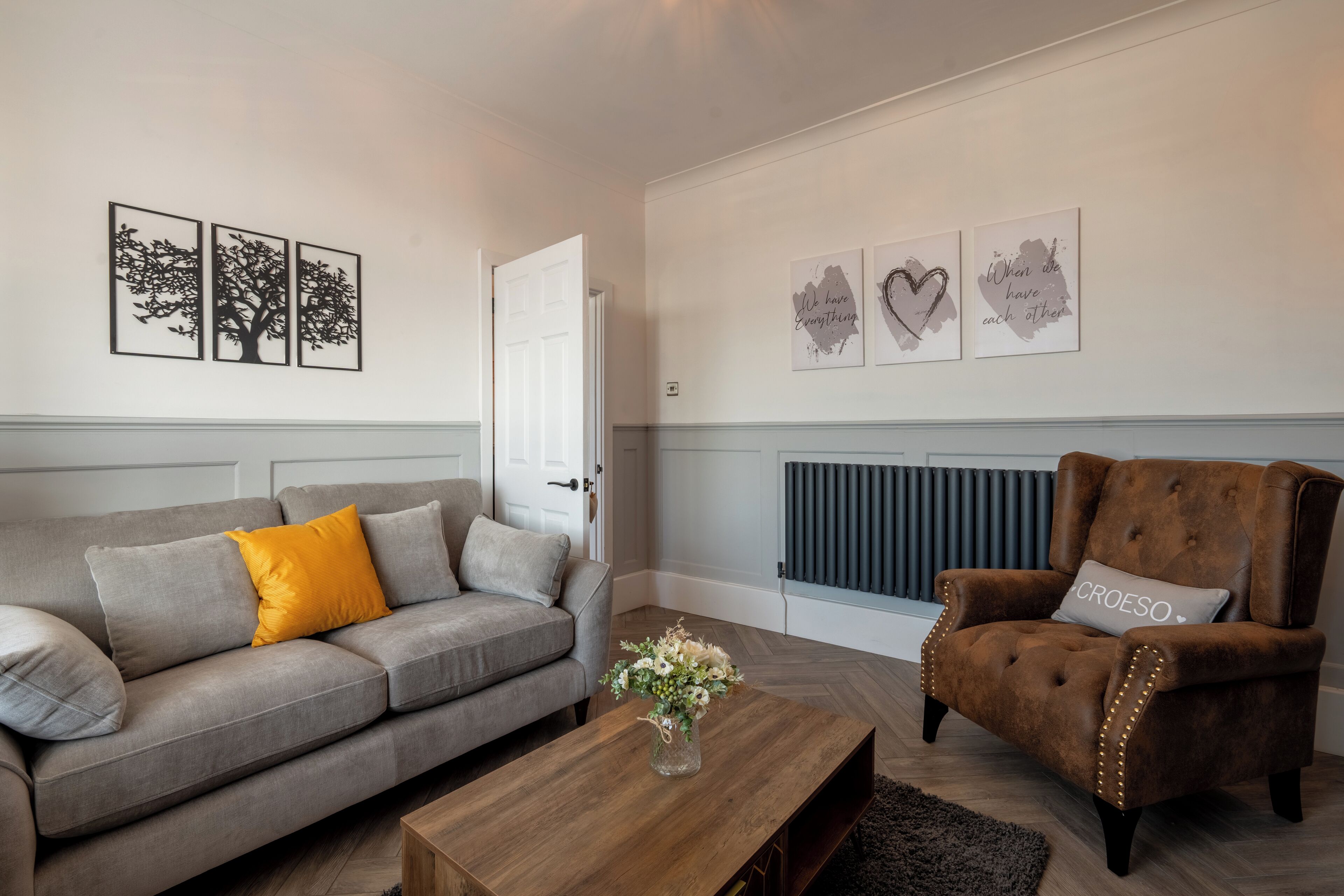Een woonkamer met een grijze bank, een bruine fauteuil en een houten bijzettafeltje. Er hangen drie kunstwerken in lijsten aan de muur en er staat een radiator onder.