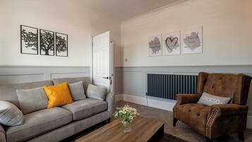 Een woonkamer met een grijze bank, een bruine fauteuil en een houten bijzettafeltje. Er hangen drie kunstwerken in lijsten aan de muur en er staat een radiator onder.
