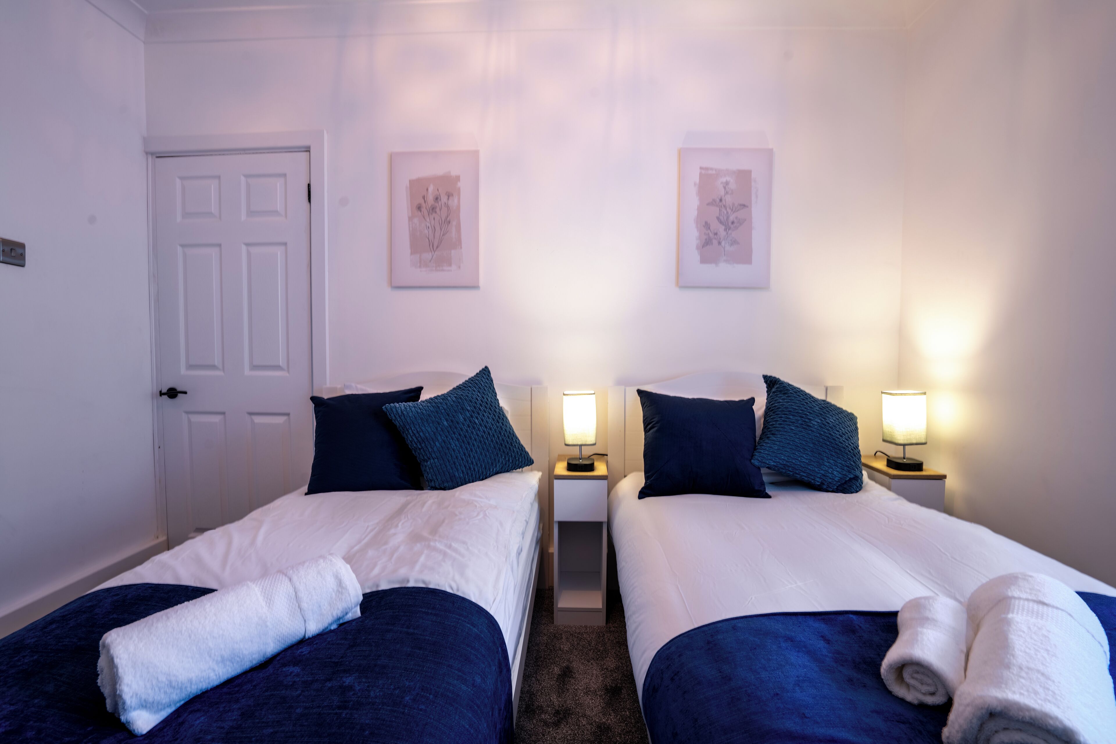A hotel room with two beds, each with a blue blanket and pillows, a white door, and two bedside lamps.