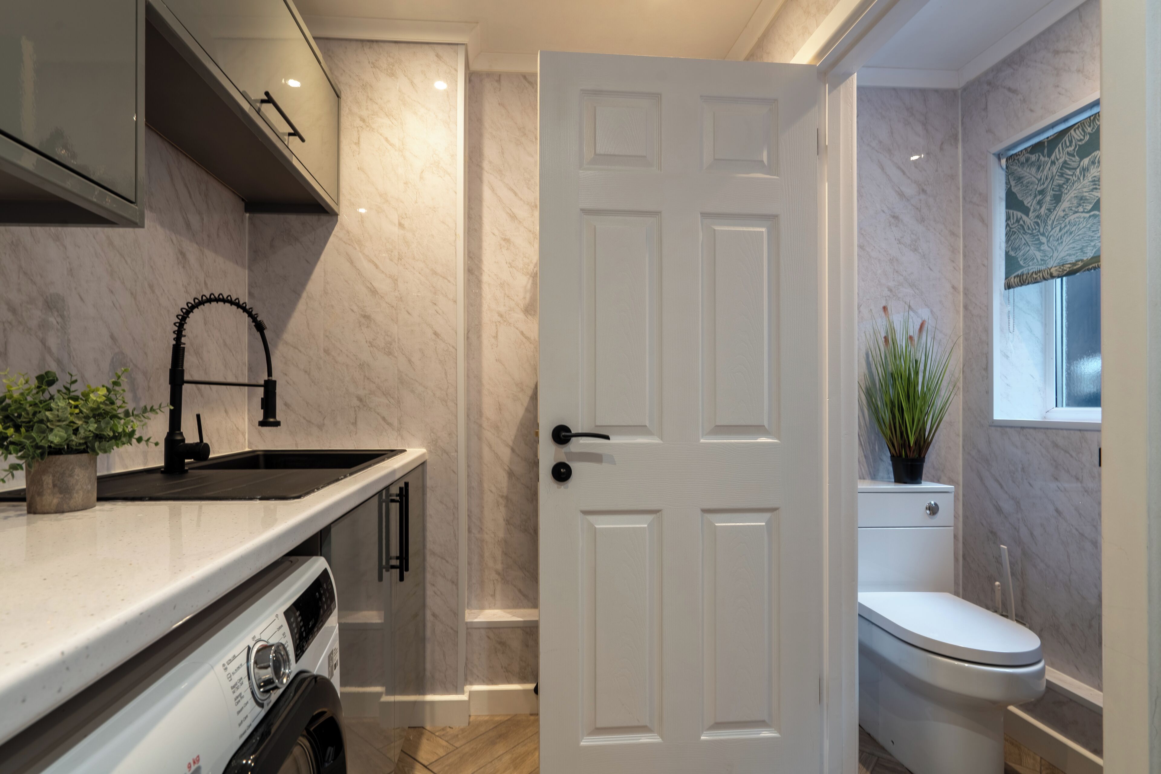Une cuisine compacte équipée d’un lave-linge encastré, d’un évier et d’une porte blanche donnant sur une salle de bain avec des toilettes et une fenêtre.