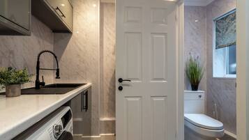 A compact kitchen with a built-in washing machine, a sink, and a white door leading to a bathroom with a toilet and a window.