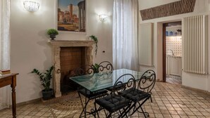 Un comedor con mesa de vidrio y sillas metálicas, una chimenea, un cuadro de un edificio y una cocina visible a través de un vano.