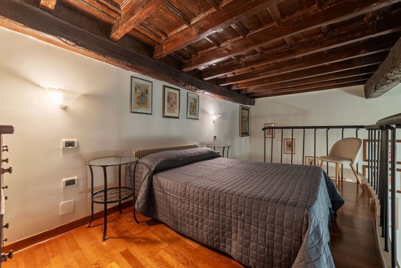 Ein Schlafzimmer mit Holzbalkendecke, einem Bett mit grauer Tagesdecke, einem kleinen runden Tisch und einem Stuhl.