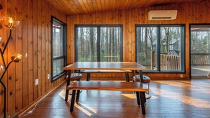 En inredning med träpanel, ett stort matbord och bänkar, stora fönster med utsikt över skogen och en luftkonditioneringsanläggning.