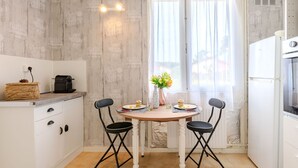 A compact kitchen with white cabinets, a small dining table with two chairs, a coffee machine, and a window with curtains. - Les Trounques by Interhome (Mimizan)