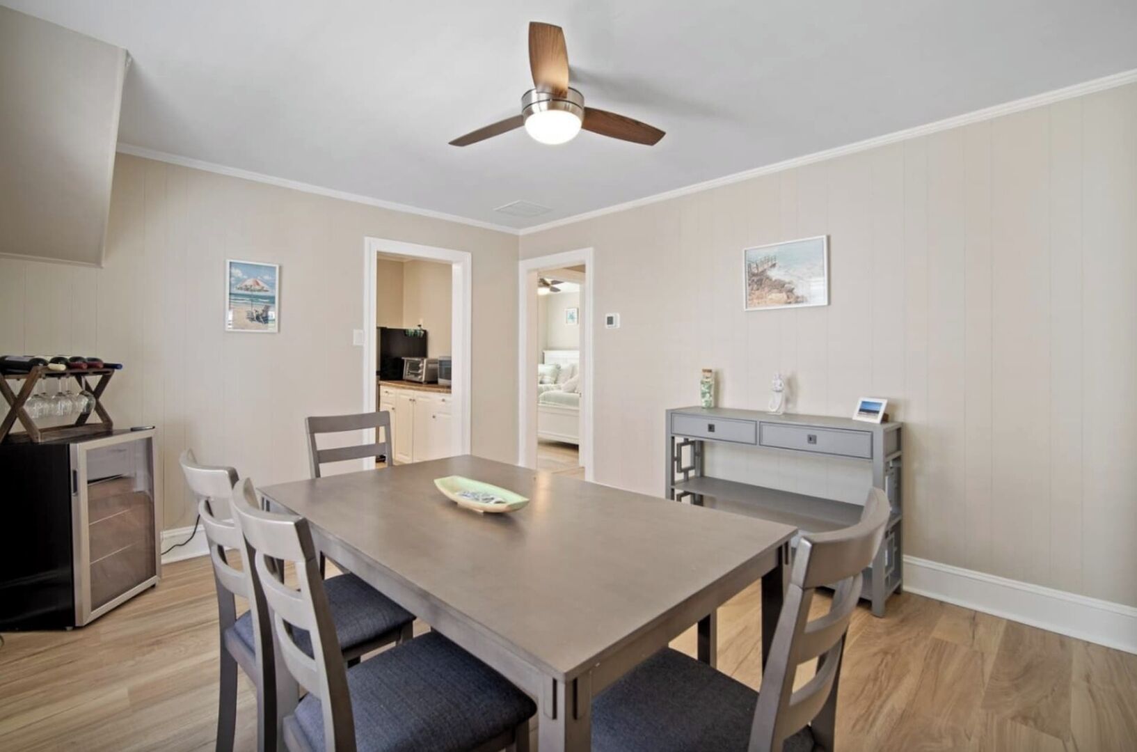 A dining area with a wooden table, four chairs, a ceiling fan, and a kitchen in the background.