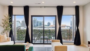 A modern living room with a sofa, a coffee table, and a view of a cityscape through large windows with black curtains.