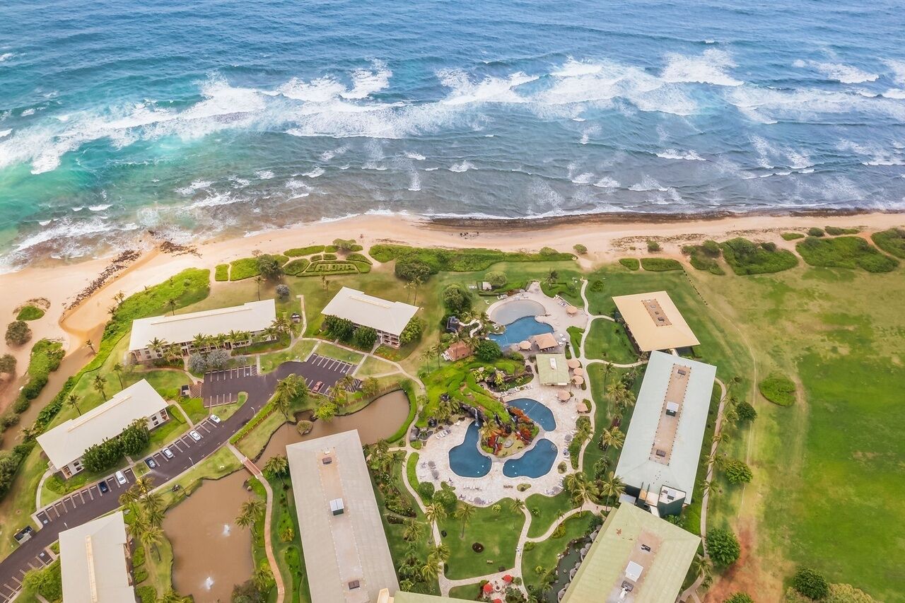 Vista aérea de un complejo turístico costero con varias construcciones, una playa y un área ajardinada con piscinas y vegetación.