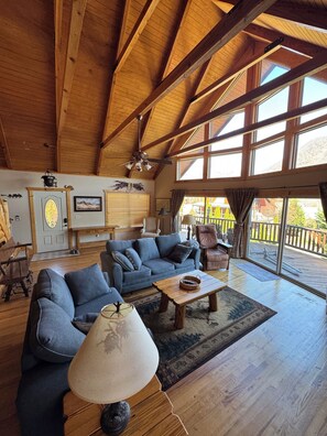 A spacious living room with a high ceiling, wooden beams, a large sofa, a coffee table, and a view of the outdoors through large windows.