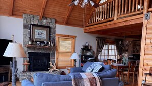 A cozy log cabin interior with a stone fireplace, a blue sofa, wooden flooring, and a dining area with wooden furniture.