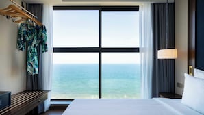 Ein Hotelzimmer mit einem großen Bett, einer hölzernen Kommode, einem aufgehängten Bademantel und Meerblick durch ein großes Fenster.