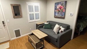 A living room with a grey sofa, a wooden coffee table, a window with blinds, a painting on the wall, and a door leading to another room. - Clean Penthouse with Balcony (Burbank)