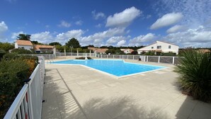 Une piscine avec une terrasse carrelée, entourée d’une clôture blanche, et des maisons en arrière-plan.