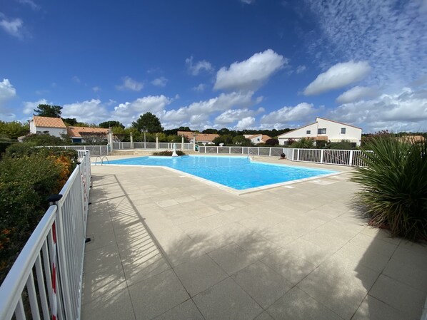 Une piscine avec une terrasse carrelée, entourée d’une clôture blanche, et des maisons en arrière-plan.