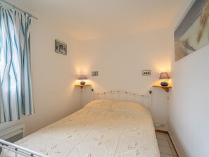 Une petite chambre à coucher, bien rangée, avec un lit simple, deux lampes fixées au mur et une fenêtre avec des rideaux.