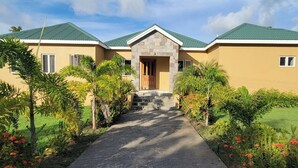 Una casa de dos pisos con entrada de piedra, techo verde y jardín con plantas y flores tropicales.
