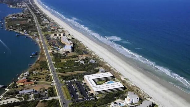 An aerial view of a coastal area with a long beachfront, a road parallel to the beach, and several buildings including a large modern structure. - Pelican View 3 Bedroom Ocean House 308 (Saint Augsutine)