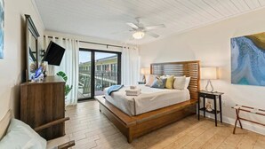A bedroom with a large bed, a ceiling fan, a TV, a wooden dresser, a nightstand, and a painting on the wall. - Pelican View 3 Bedroom Ocean House 308 (Saint Augsutine)