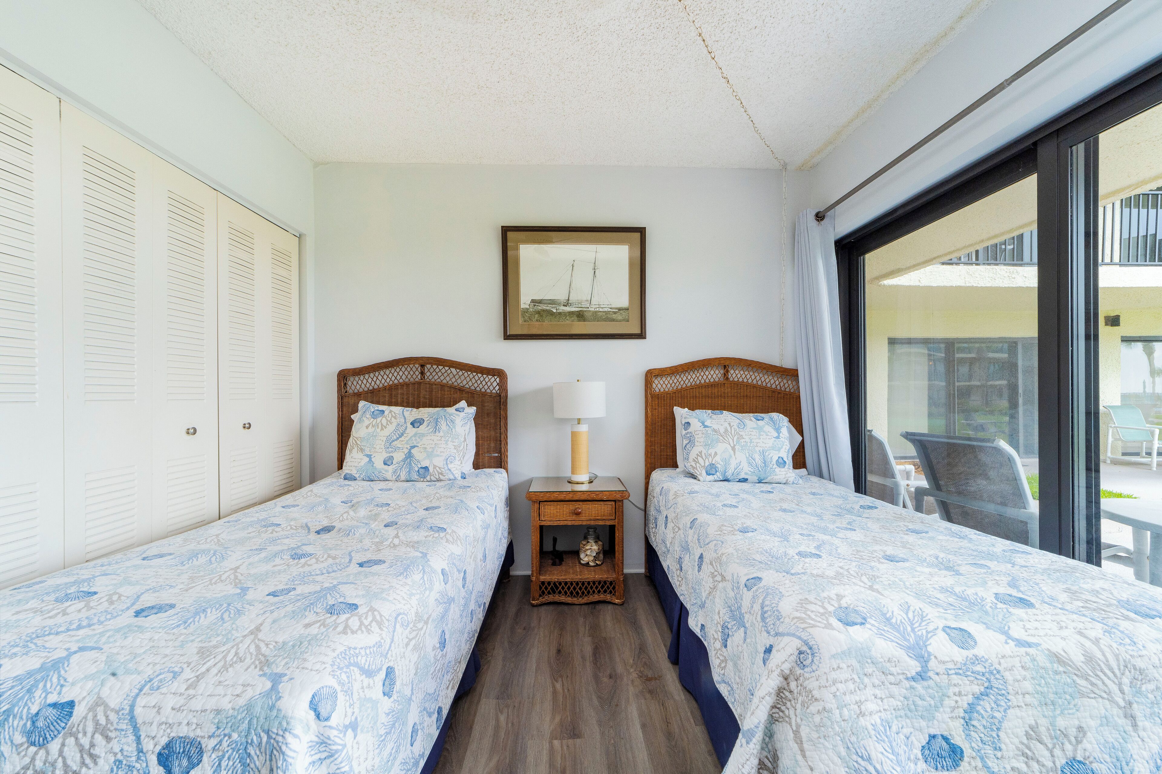 Et hotelværelse med to senge, et lille bord med en lampe, et billede i ramme på væggen og udsigt til den udendørs balkon.