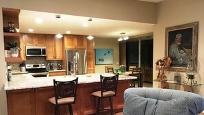 A modern kitchen with wooden cabinets, a central island with bar stools, a refrigerator, and a microwave. - SANDPIPER.....Breathtaking Views of Mountains and Pool! (Palm Desert)