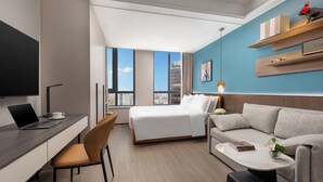 Ein modernes Hotelzimmer mit einem Bett, einem Schreibtisch mit Laptop, einer Couch und einem Fernseher.