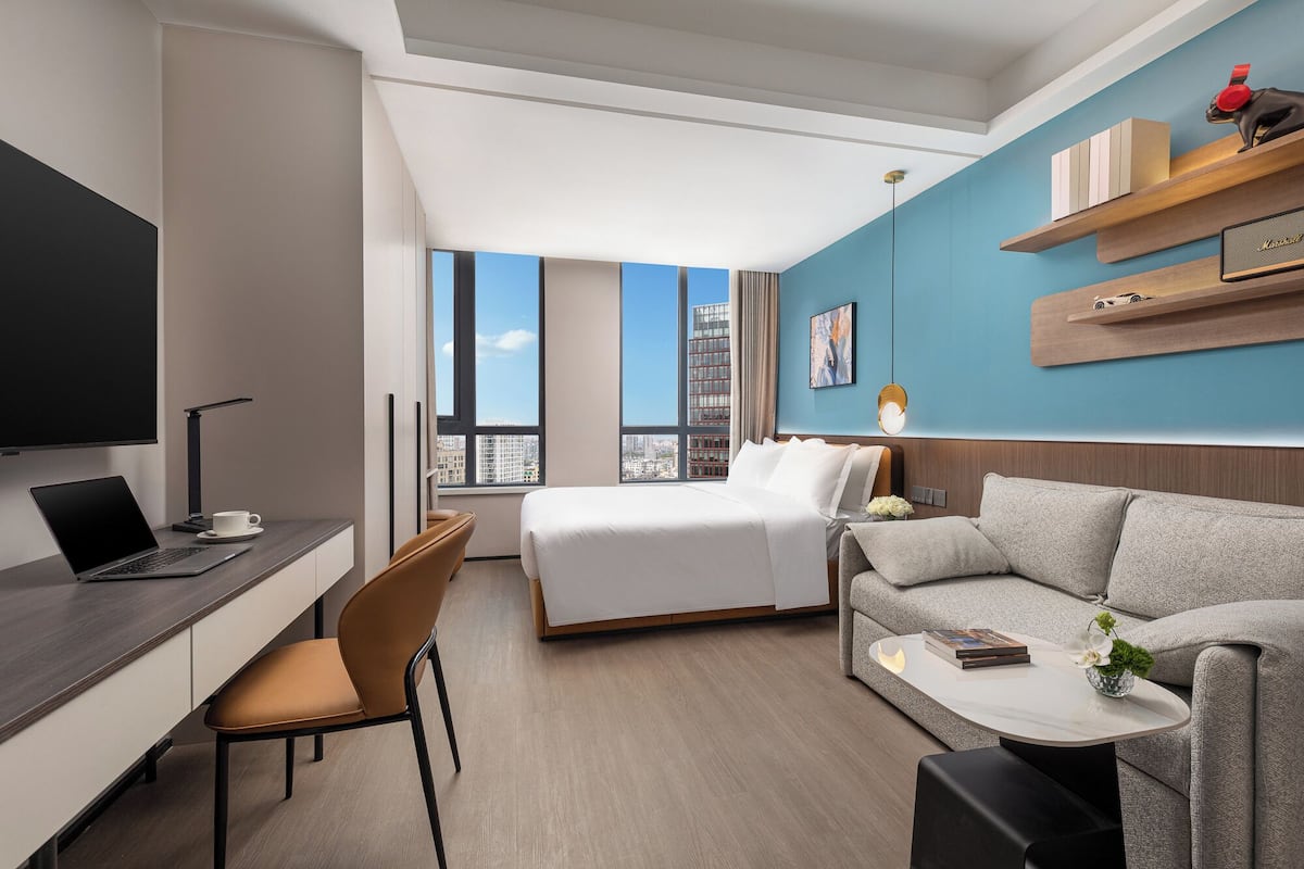 A modern hotel room with a bed, a desk with a laptop, a sofa, and a TV.
