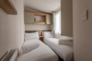A bedroom with two beds, a small shelf with a book, a window with curtains, and a radiator.