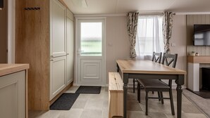 A compact kitchen with a wooden cabinet, a dining table with chairs, a black bench, and a television on the wall.
