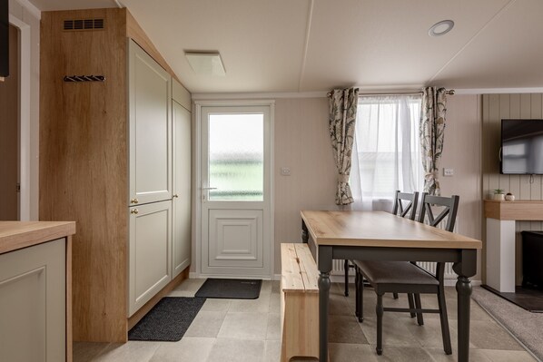 A compact kitchen with a wooden cabinet, a dining table with chairs, a black bench, and a television on the wall.