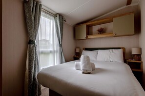 A neatly made bed with white linens and two towels, a bedside table with a lamp, a nightstand with a lamp, a wooden headboard, a wall-mounted shelf with a vase, and a window with curtains.