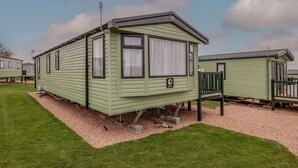 A green mobile home with a small porch, a window with curtains, and a satellite dish.