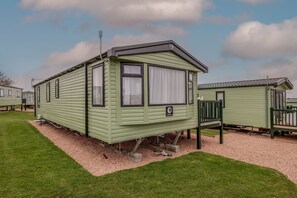 A green mobile home with a small porch, a window with curtains, and a satellite dish.