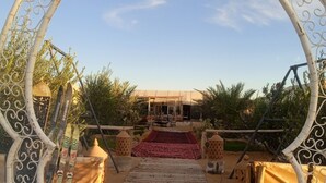 An outdoor pathway leading to a covered area with a red carpet, surrounded by decorative metal arches and palm trees. - Sahara Merzouga Camp (Hassilabied)