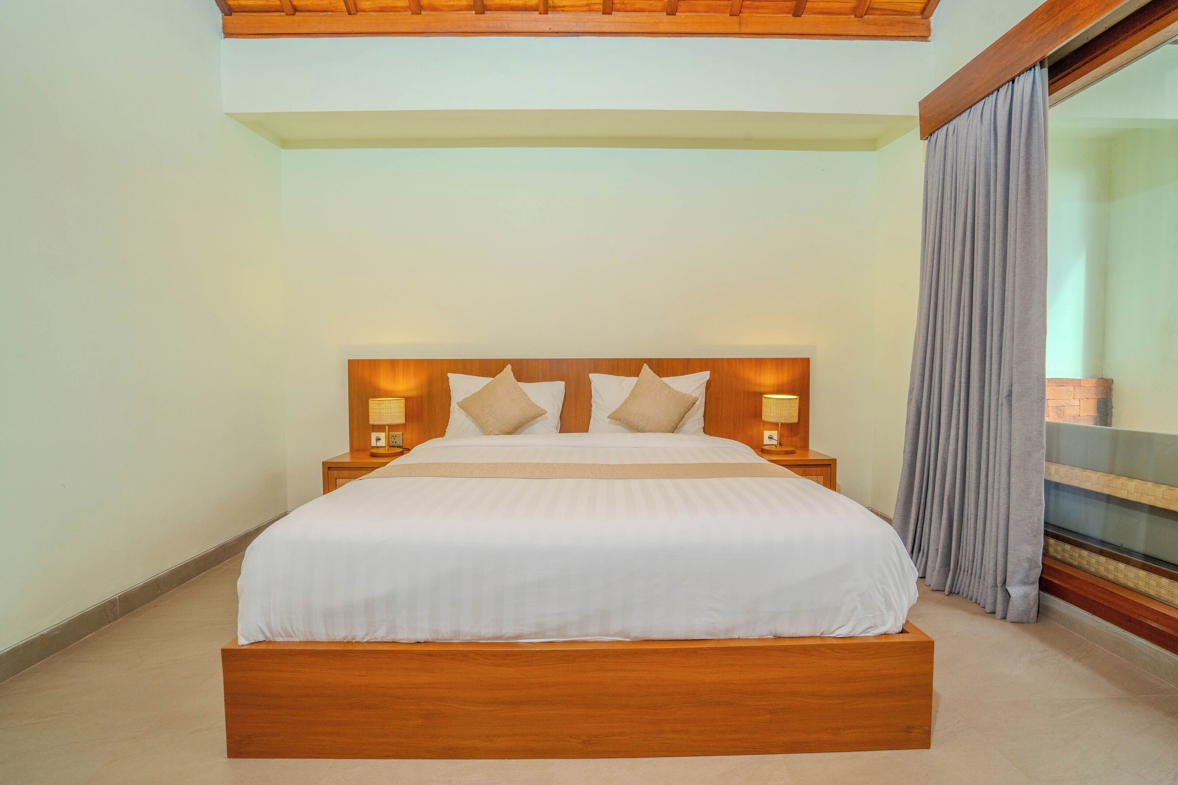A large bed with a wooden headboard, two bedside tables with lamps, and a view of a balcony with curtains.