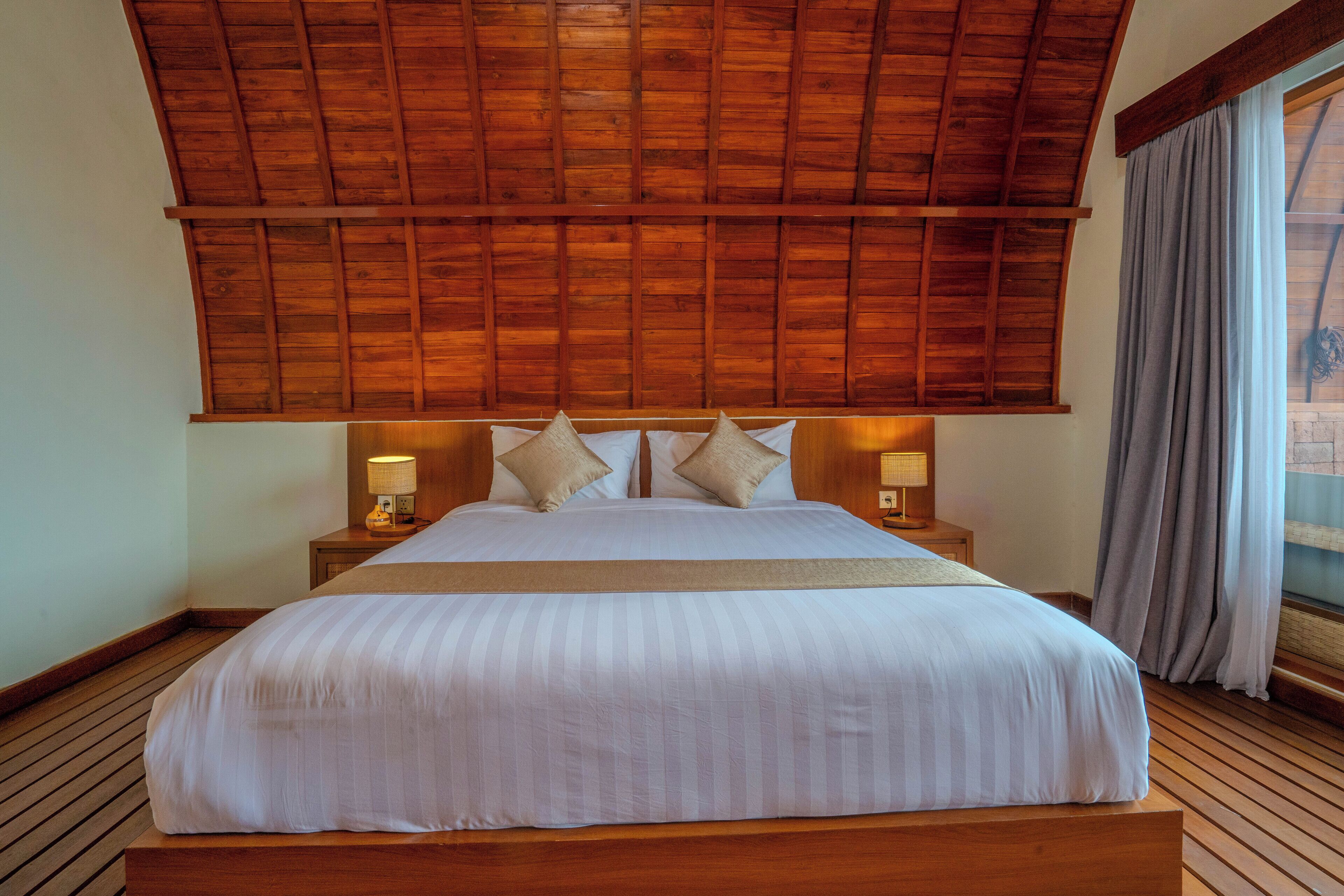 A large bed with a wooden headboard, two bedside lamps, and a view of the outdoors through a window with curtains.