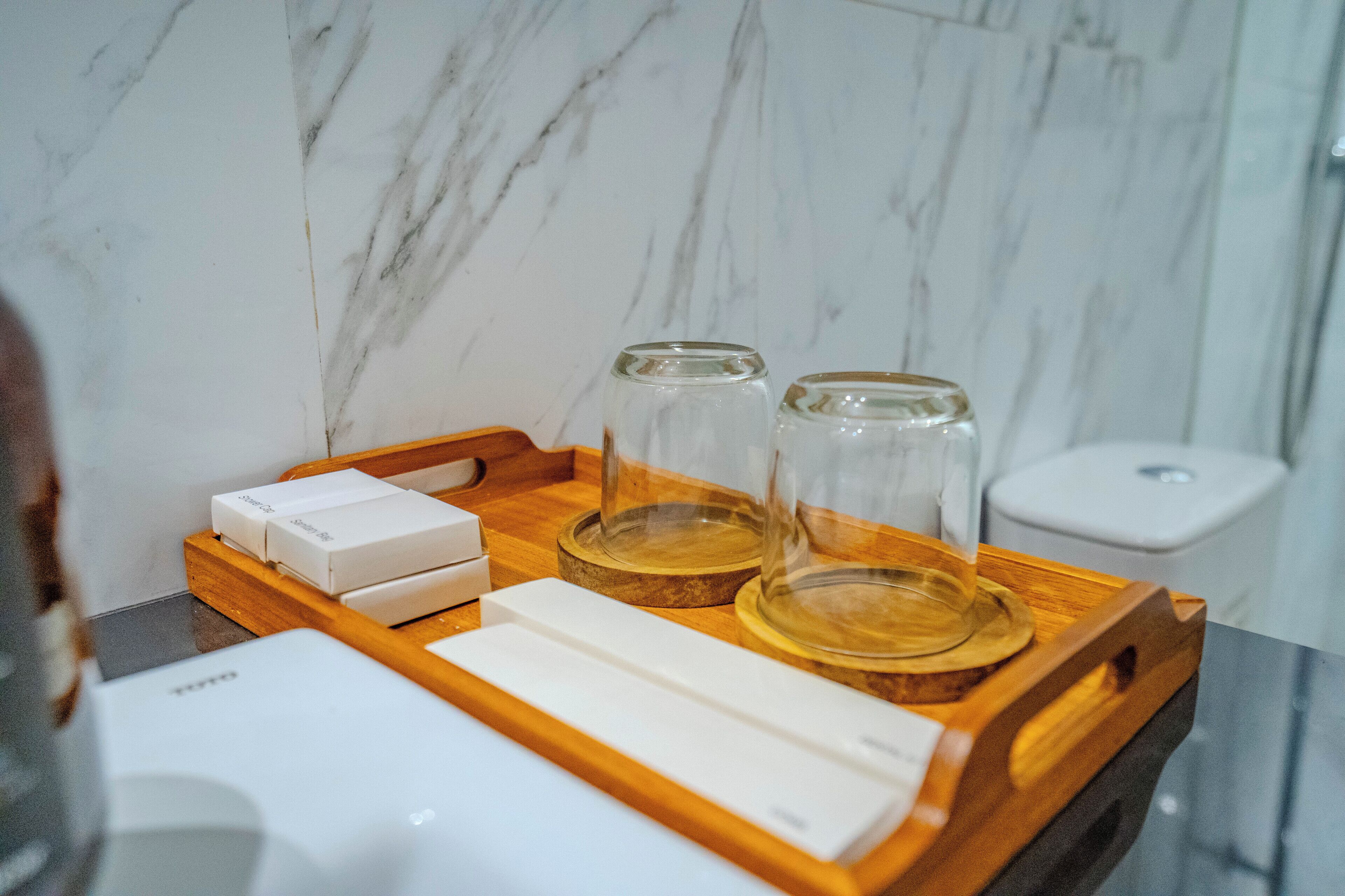 Una bandeja de madera con una caja blanca, dos recipientes de vidrio y un objeto rectangular blanco sobre una encimera de mármol.