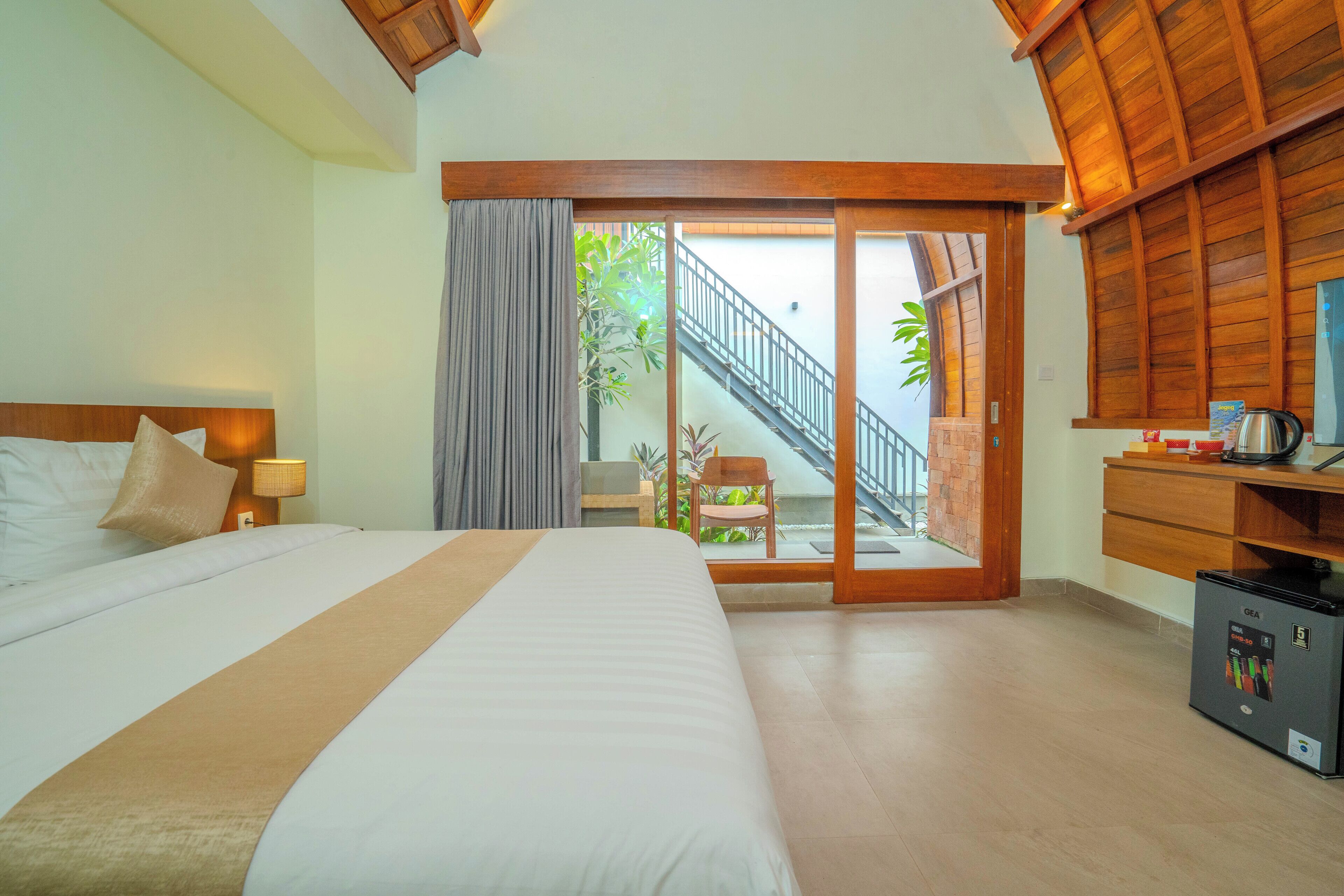 A hotel room with a large bed, a desk with a kettle, and a view of a staircase leading to an outdoor area.
