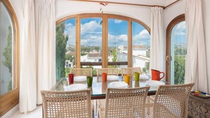 A dining area with a glass table and wicker chairs, large windows with curtains, and a view of buildings and greenery.