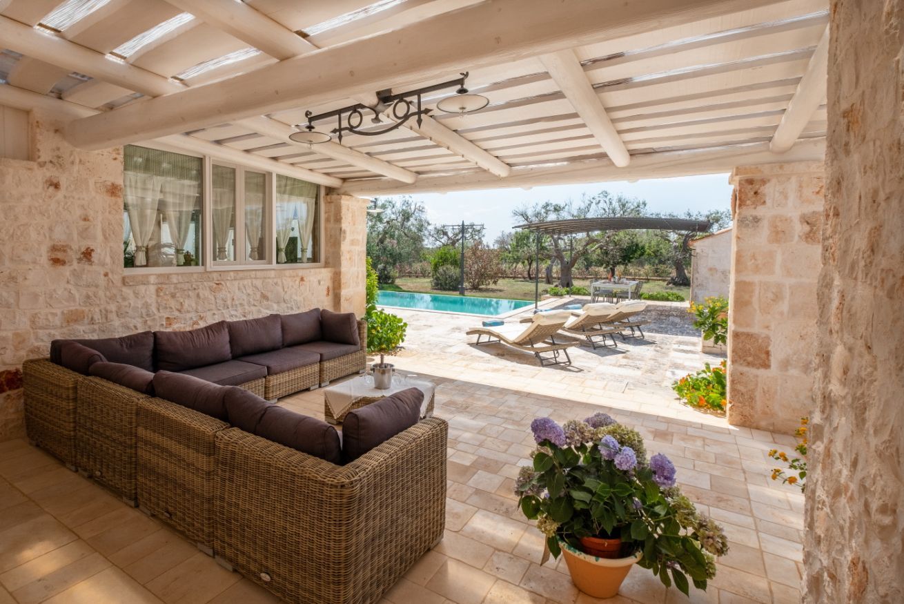 A covered outdoor seating area with wicker furniture, a pool, and a garden view.
