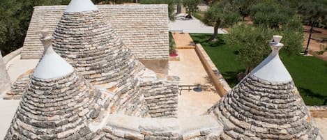 A traditional stone-roofed building with multiple conical structures, surrounded by a landscaped garden and olive trees.
