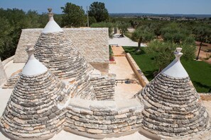A traditional stone-roofed building with multiple conical structures, surrounded by a landscaped garden and olive trees.