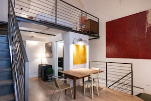 A loft-style apartment with a staircase, a dining area with a wooden table and chairs, a red abstract painting, and a loft bed.