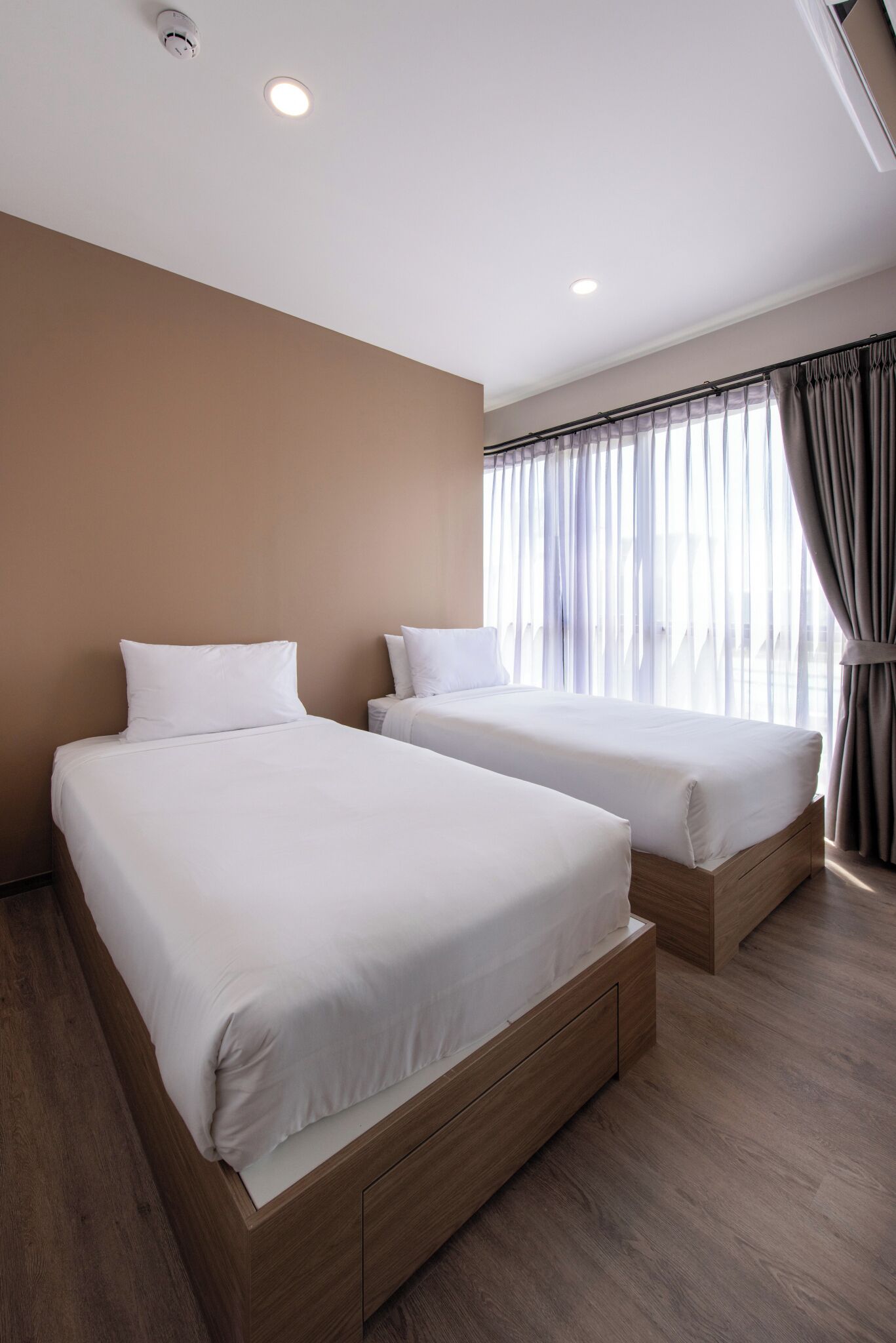 A hotel room with two beds, wooden flooring, a large window with curtains, and a ceiling with recessed lighting.