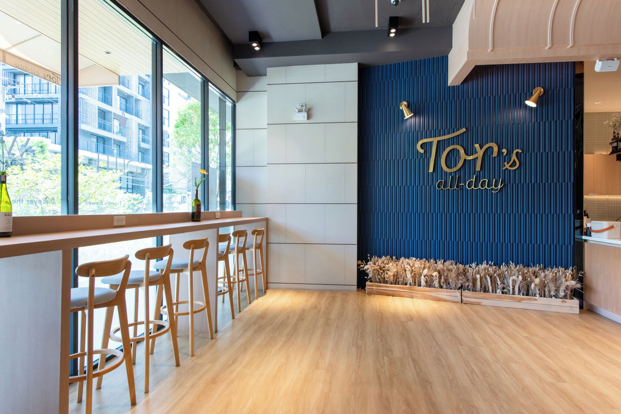 A modern bar area with wooden stools, a blue accent wall with 'Tony's all-day' signage, and a view of the outside through large windows.