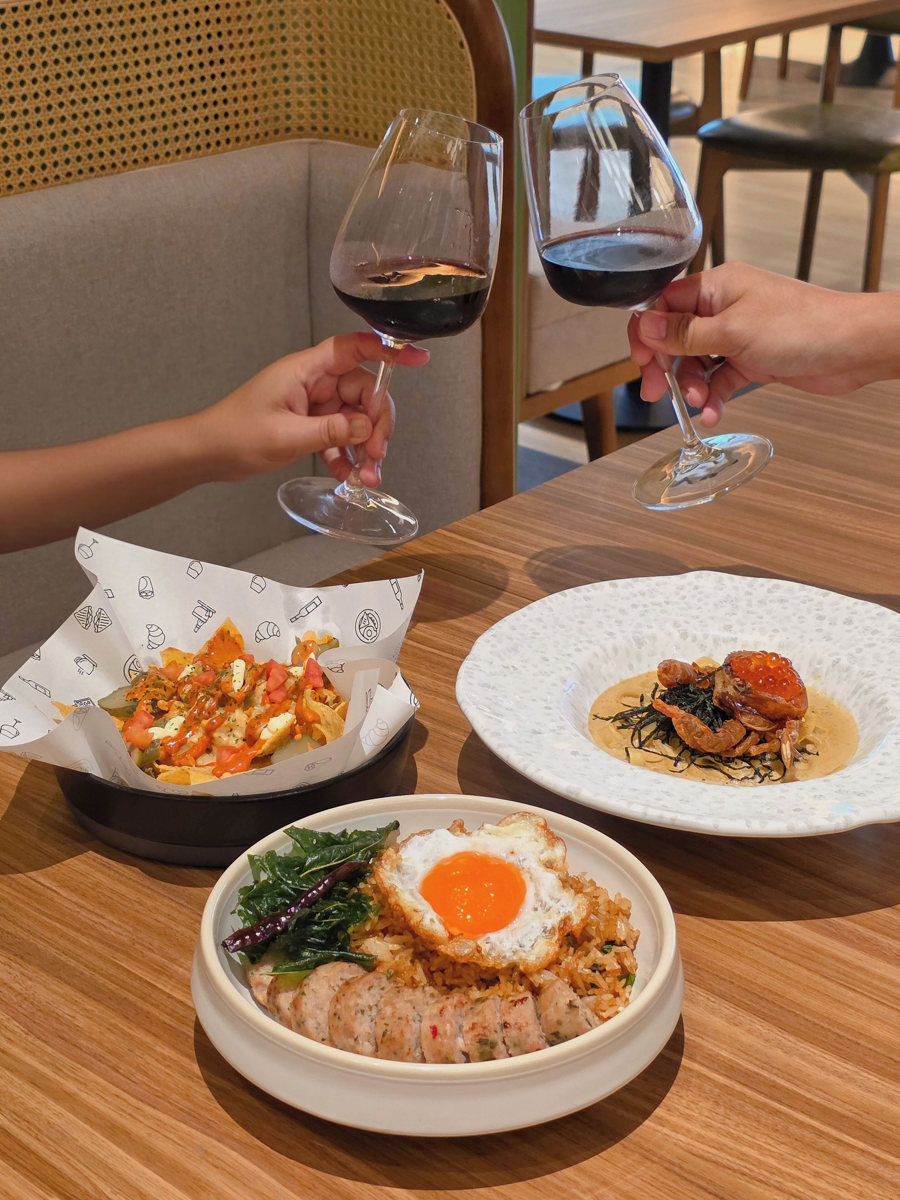A dining table with three dishes: nachos, seafood with sauce, and fried rice with egg. Two people are holding wine glasses.