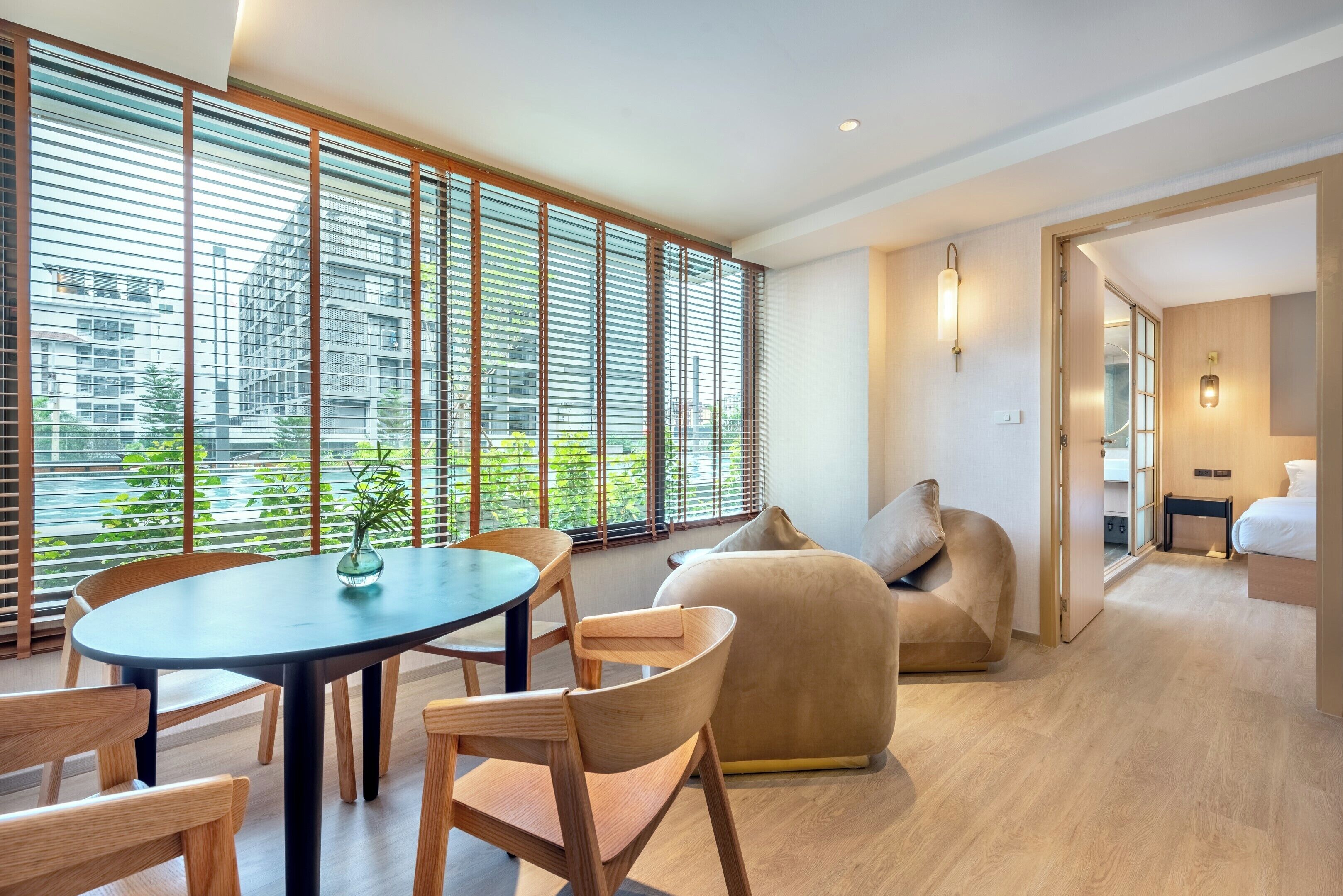 A modern interior with a round table, wooden chairs, a beige sofa, and large windows with blinds.