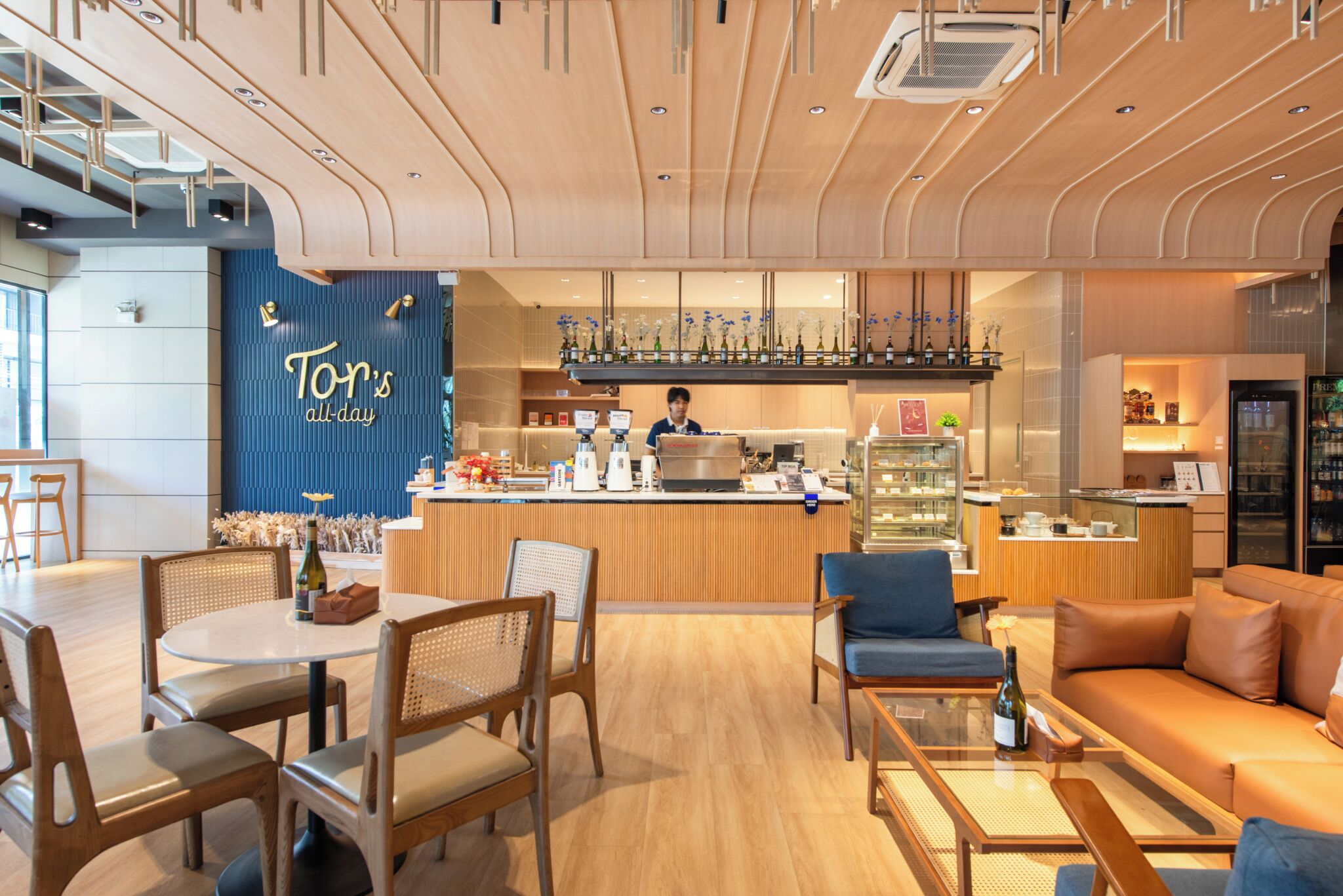 A modern hotel lobby with a bar area, seating arrangements, and a visible kitchen in the background.