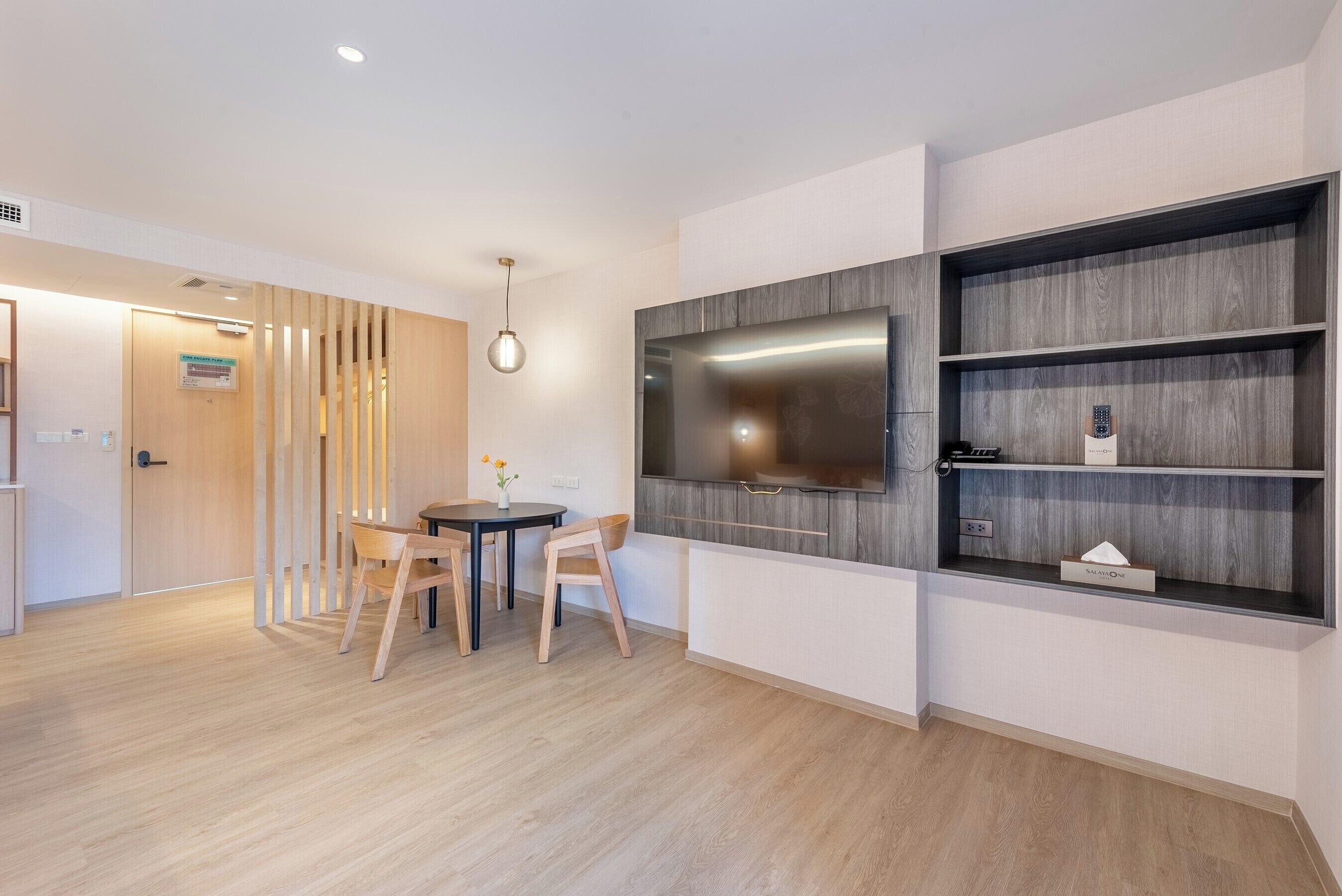 A modern living room with a flat-screen TV mounted on a wooden wall, a dining area with a round table and chairs, and a small shelf with a tissue box.